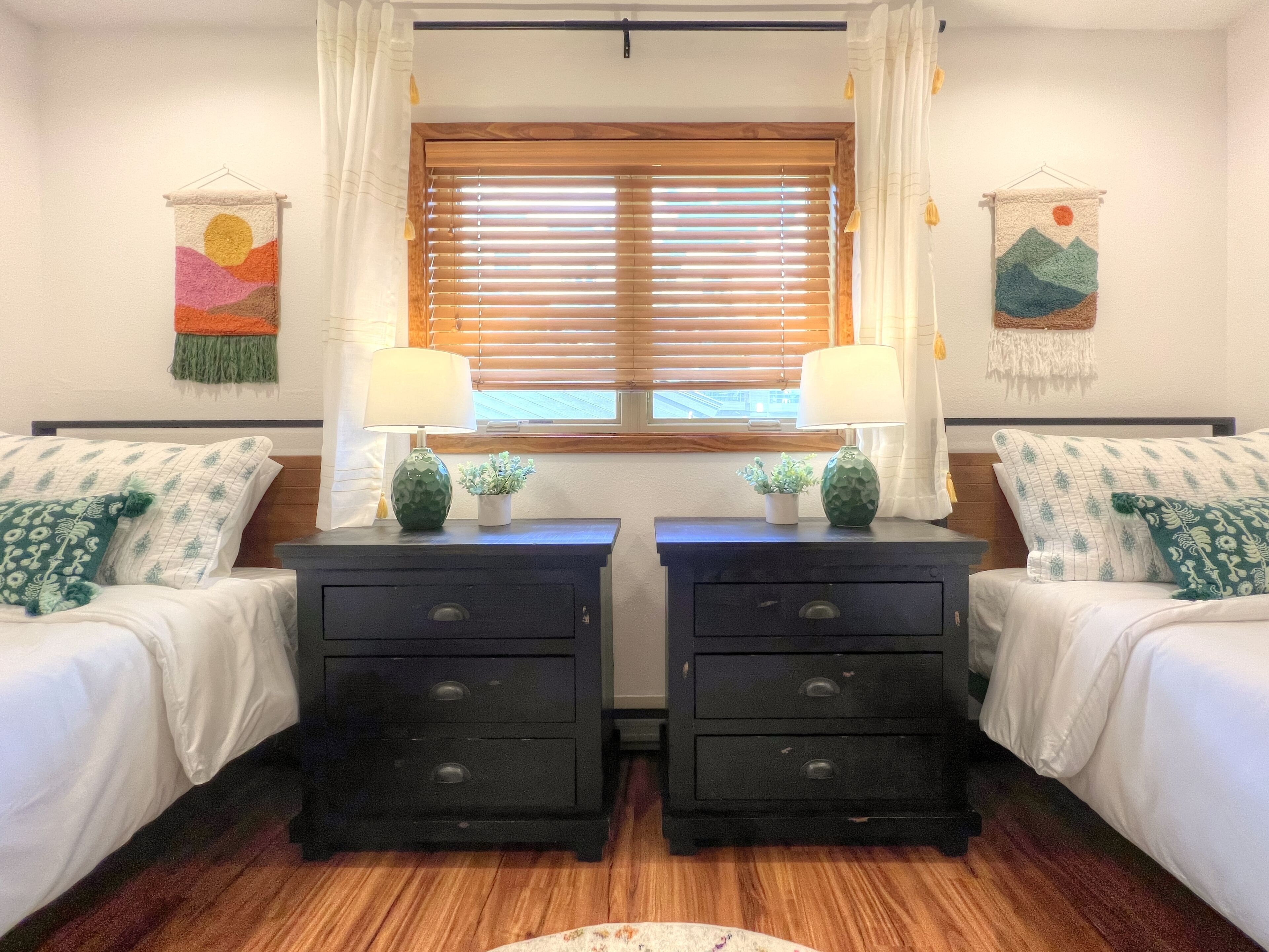 Ein Zimmer mit zwei Betten, jeweils mit einem Nachttisch, einer Lampe und einer kleinen Pflanze. Es gibt zwei Wanddekorationen, je eine an jeder Seite des Fensters.