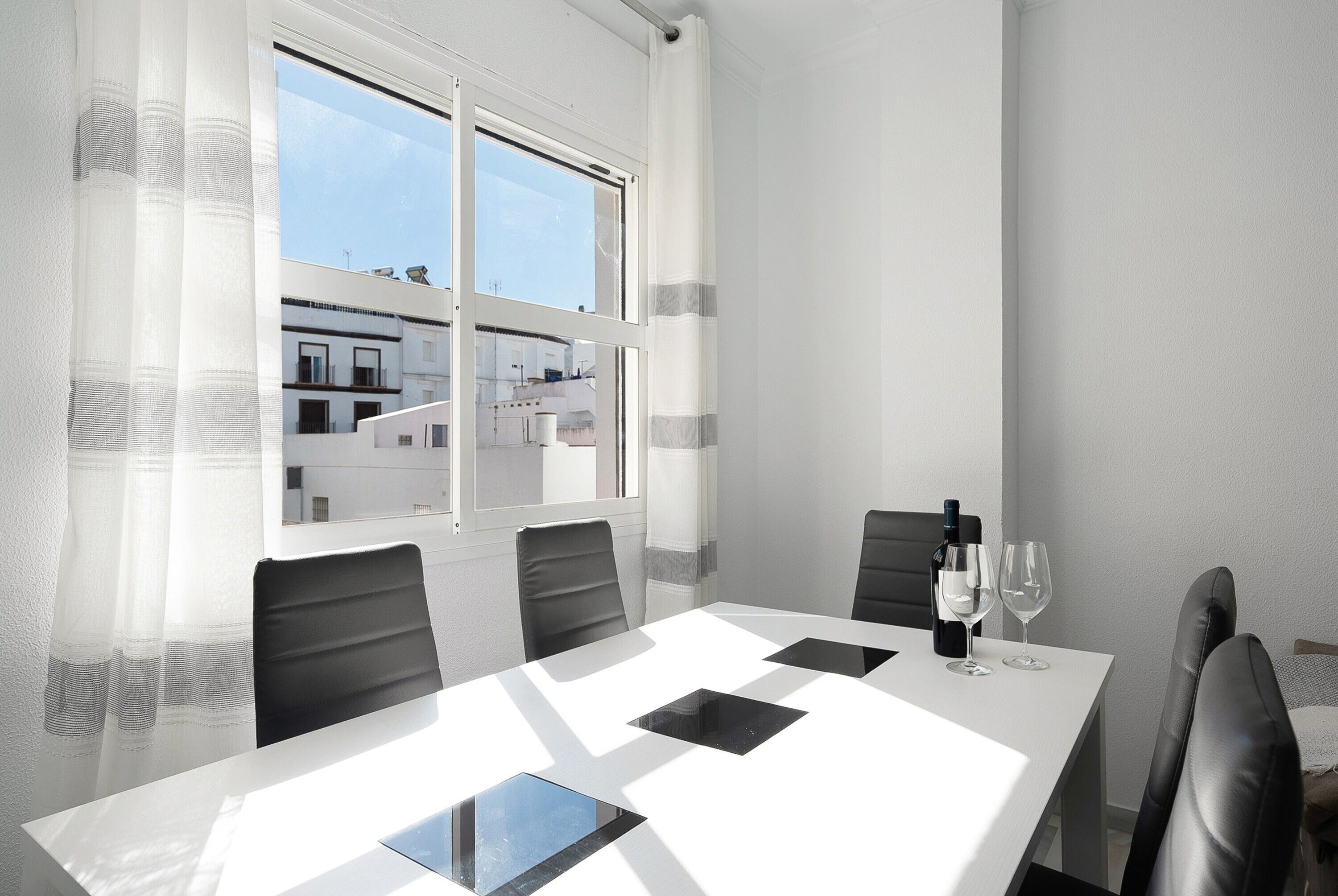 A modern dining room with a white table, black chairs, and a view of buildings through a window.