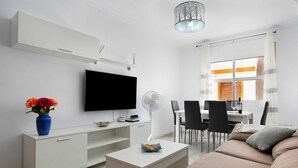 A modern living room with a flat-screen TV, white furniture, a dining area with chairs, and a large window with curtains. - Apartment 'Romo' close to the Beach with Balcony & Wi-Fi (conil de la frontera)