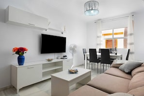 Un salon moderne doté d’un téléviseur à écran plat, de meubles blancs, d’un coin repas avec des chaises et d’une grande fenêtre agrémentée de rideaux.