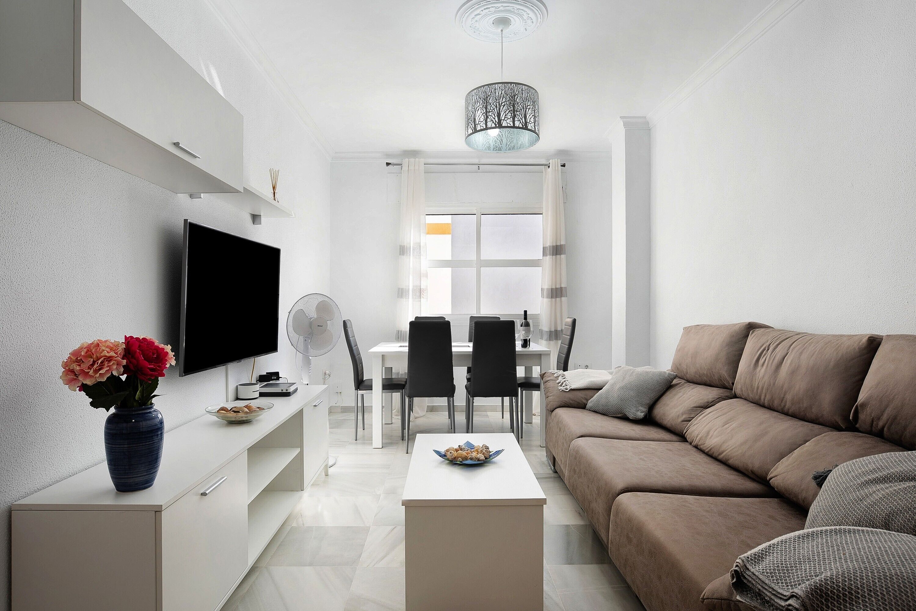 A modern living room with a flat-screen TV, a sofa, a dining area with a table and chairs, and a vase with flowers on a console table.