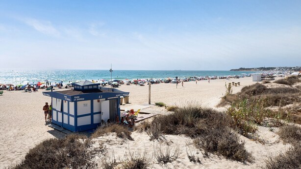 Een strand met een strandwachterpost, parasols en mensen.