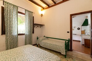 Une chambre avec deux lits, une fenêtre avec des rideaux, une étagère fixée au mur et une porte donnant sur un coin cuisine.