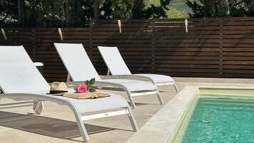 Zona de piscina con dos sillones blancos, una mesa pequeña con un libro y una rosa, y una valla de madera al fondo.