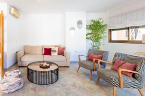 A living room with a sofa, two patterned armchairs, a coffee table, a potted plant, and a window with curtains.