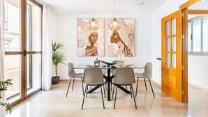 Un coin repas avec une table en verre, quatre chaises, deux lampes suspendues et des œuvres d’art accrochées au mur.