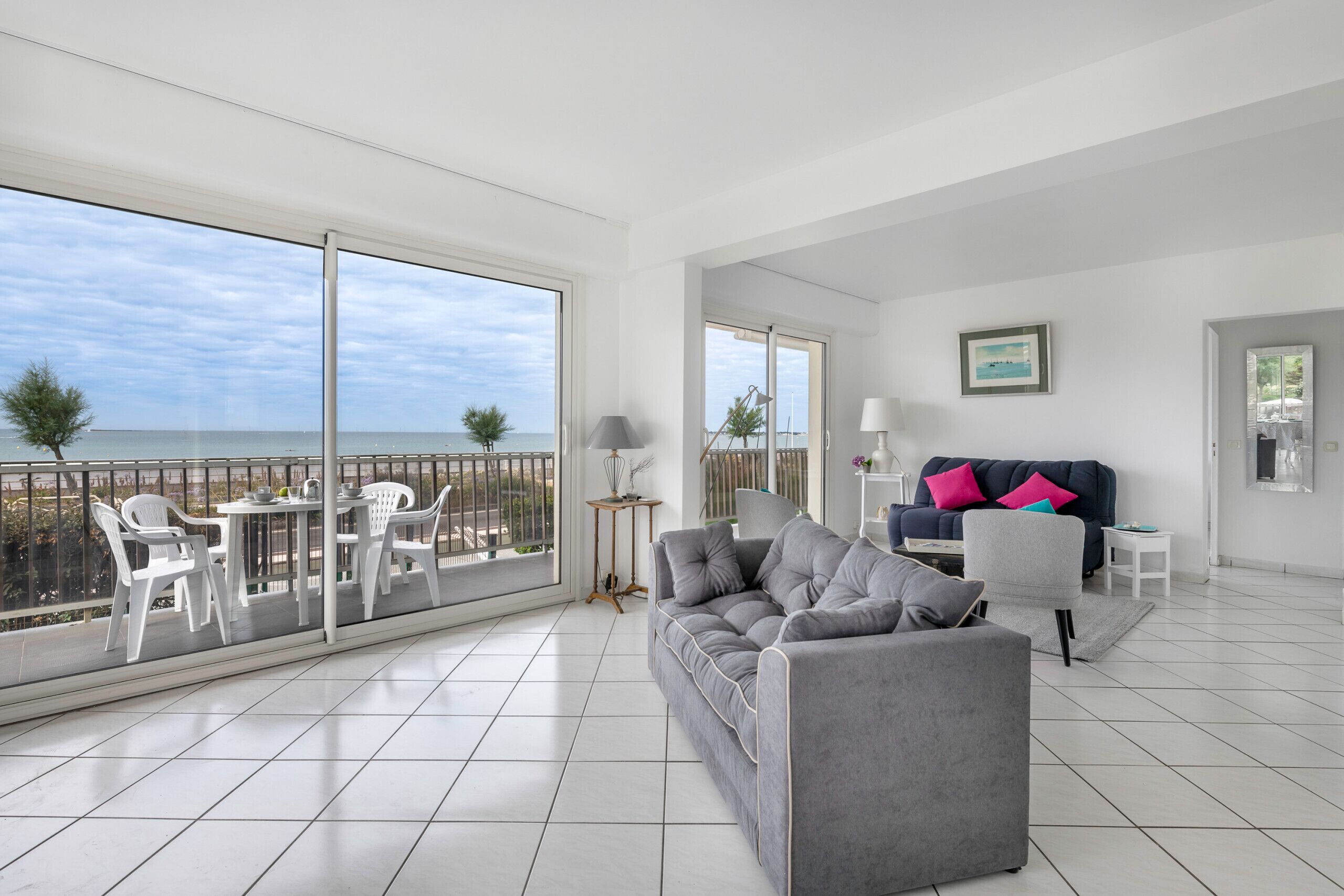 A spacious living area with a grey sofa, white chairs, and a balcony overlooking the sea.