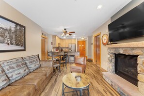 Cozy living room with a comfortable sofa, stone fireplace, and modern TV. The adjacent kitchen features wooden cabinetry and a dining table.
