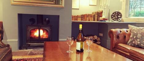 Una sala de estar acogedora con chimenea, una mesa de centro de madera con una botella de vino y copas, un sofá de cuero y una estantería con libros y un reloj.