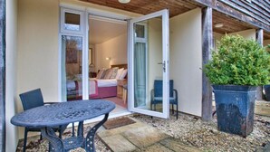 A cozy bedroom with a bed, a nightstand, and a window. There is a patio with a table and chairs, and a potted plant. - The Vines (Bampton)