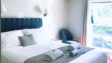 Una cama bien hecha con ropa de cama blanca y un plaid estampado, un cabecero tapizado en azul y una lámpara de noche.