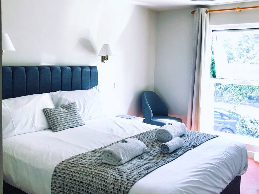 Una cama bien hecha con ropa de cama blanca y un plaid estampado, un cabecero tapizado en azul y una lámpara de noche.