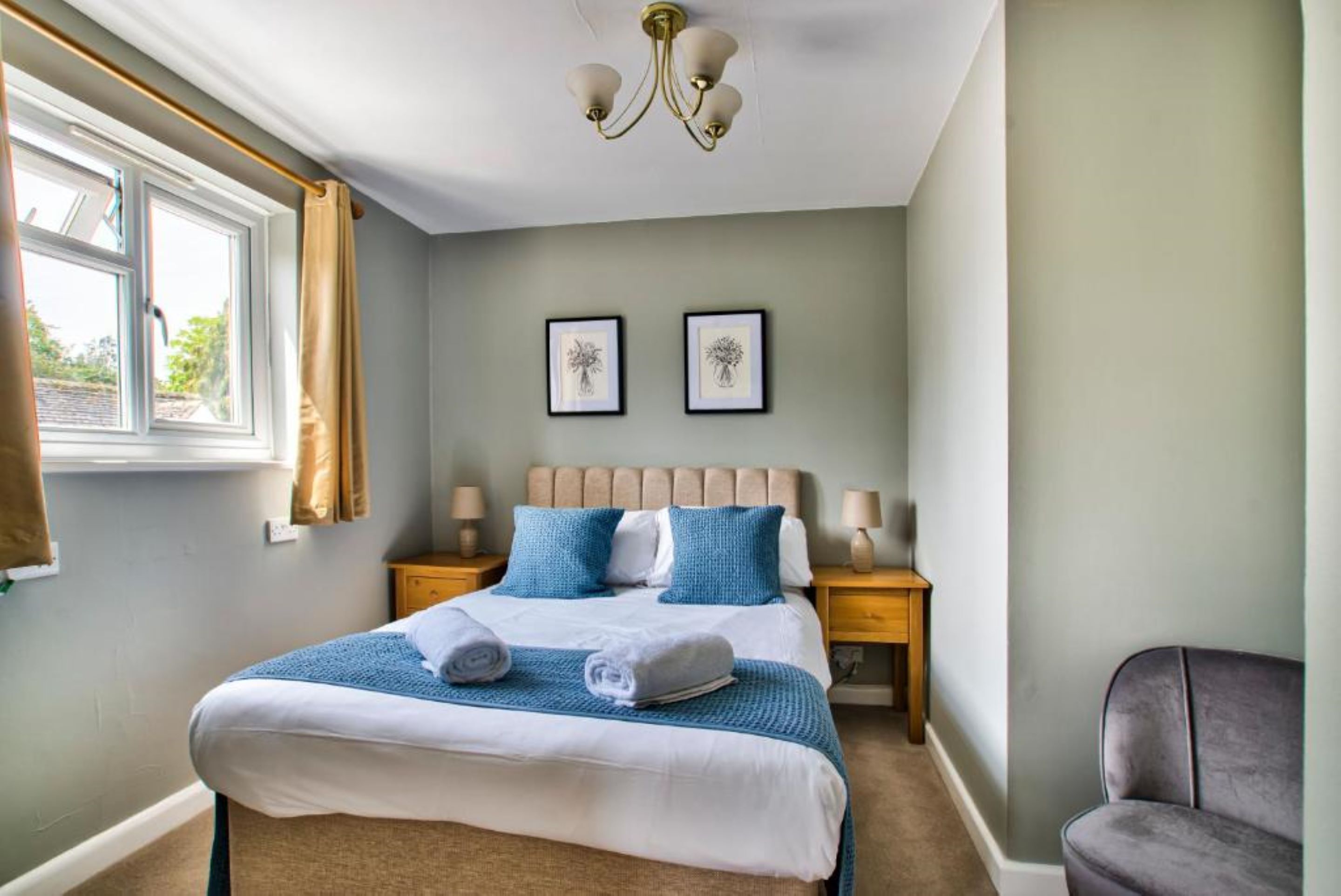 A bedroom with a bed, two bedside tables, a chandelier, a window with curtains, and two framed pictures on the wall.