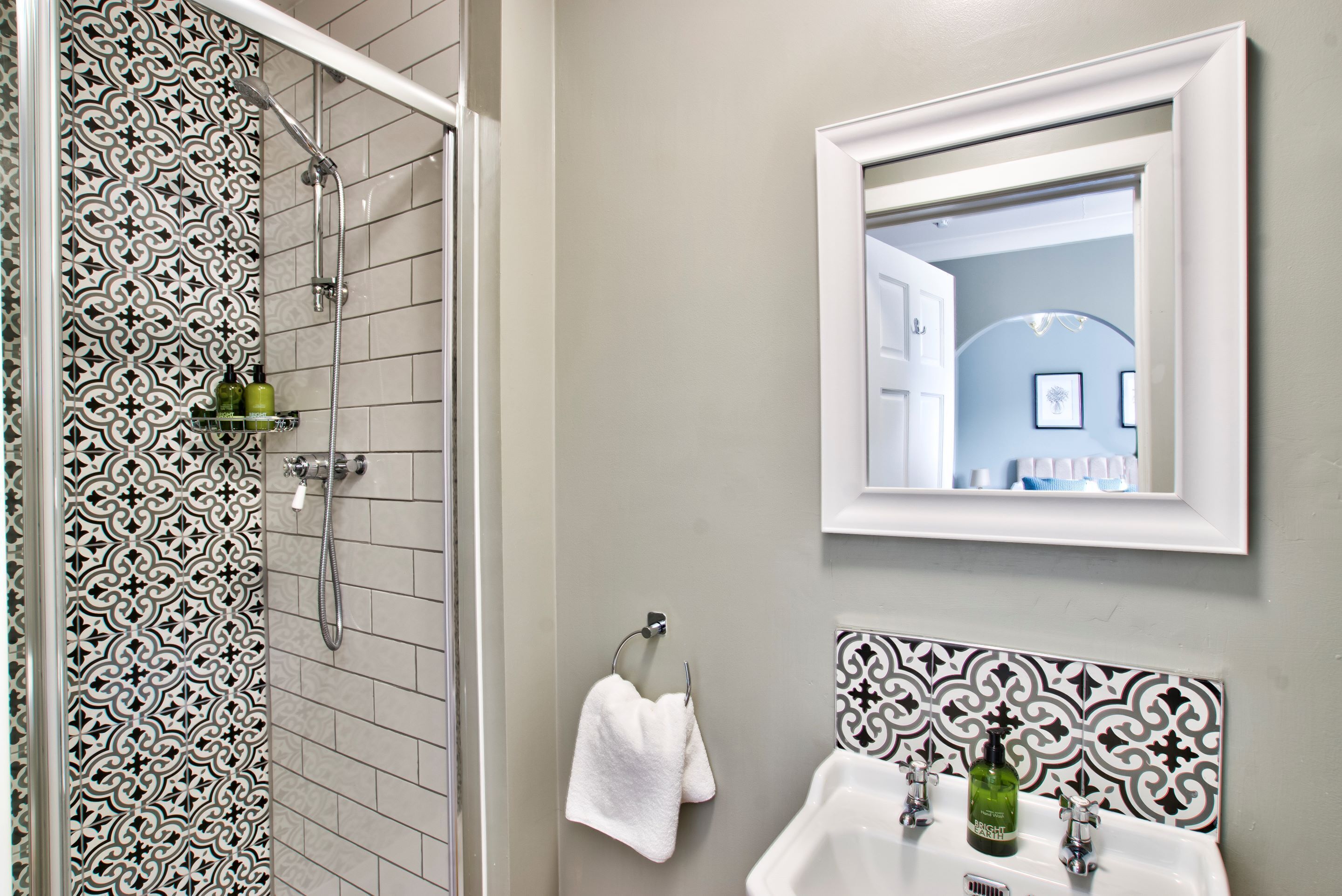 A bathroom with patterned tiles, a shower, a sink with two faucets, a mirror, and a towel rack.