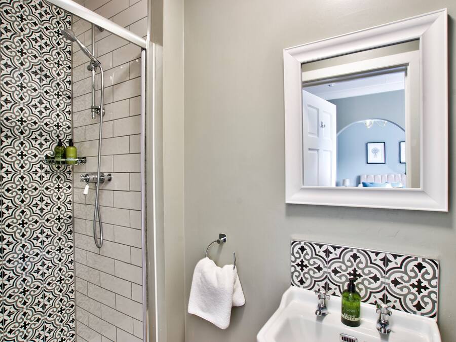 A bathroom with patterned tiles, a shower, a sink with two faucets, a mirror, and a towel rack.