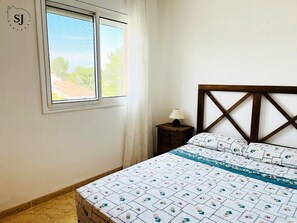 A bedroom with a bed, a nightstand, a window, and a view of the outside.
