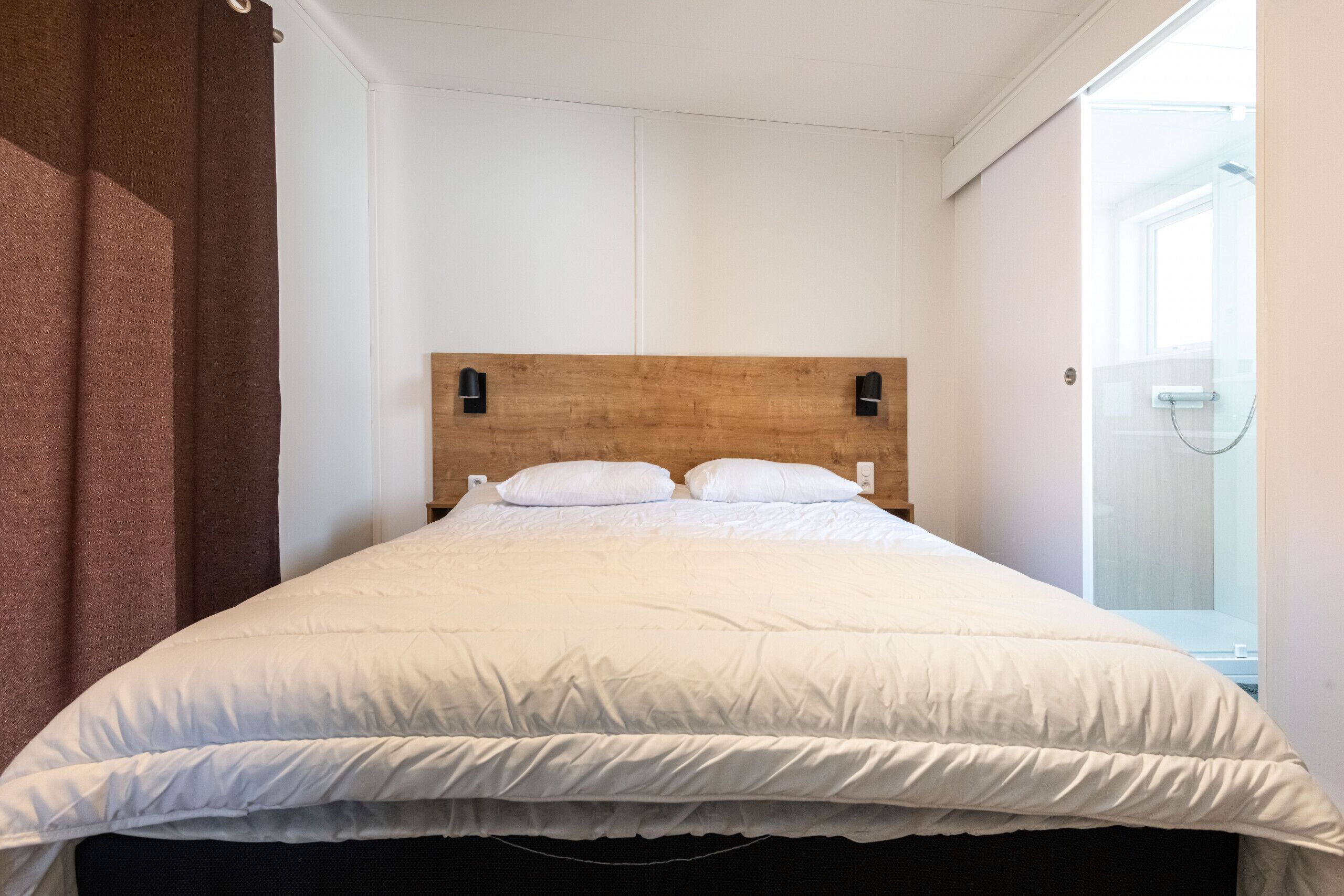 A neatly made bed with white bedding and two pillows, a wooden headboard, and a wall-mounted light fixture.