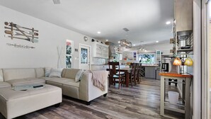 Una sala de estar moderna con un sofá seccional color beige, una zona de comedor con sillas de madera, y una cocina con armarios blancos y una lámpara colgante.