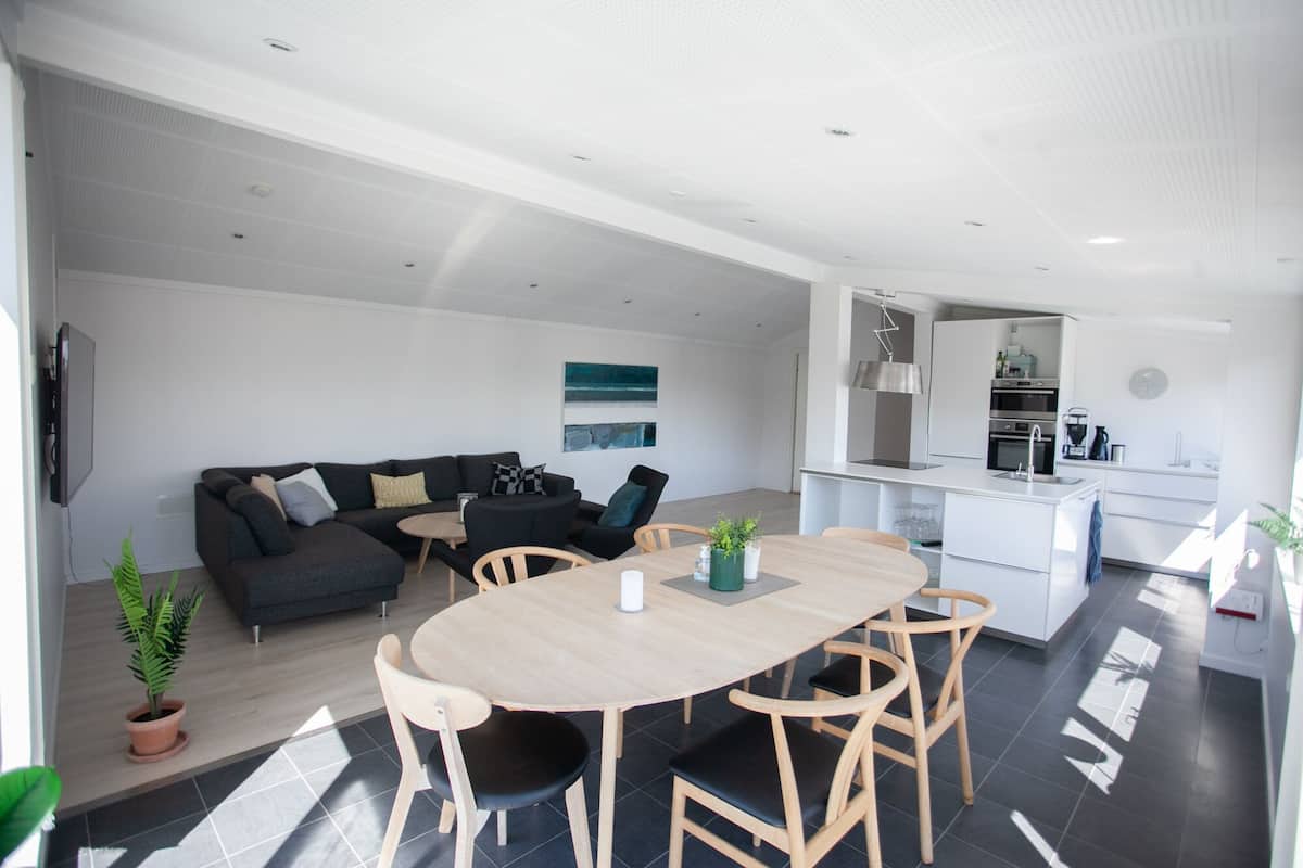 Ein moderner Wohnbereich mit einem dunkelgrauen Sofa, einem runden Holzesstisch mit Stühlen, einer Küchenzeile mit weißen Schränken und einem großen Fenster, das für natürliches Licht sorgt.