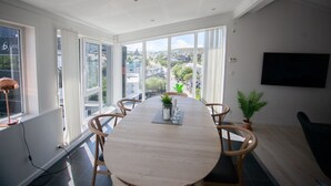 A spacious dining area with a large table and chairs, large windows offering a city view, a mounted TV, and a potted plant.