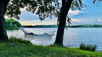 Un hamac est tendu entre deux arbres, au bord d’un lac, offrant une vue dégagée sur l’eau et les arbres qui se dressent au loin.