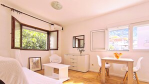 Een kamer met een bed, een bureau, een stoel, een klein tafeltje met fruit en een raam met uitzicht op groen en gebouwen.