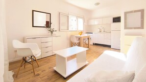 Een moderne, goed verlichte woonkamer met een witte bank, een klein wit tafeltje, en een eethoek met een houten tafel en stoelen.