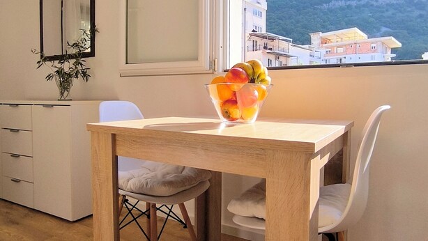Een moderne eetruimte met een houten tafel, witte stoelen, een schaal fruit, een witte kast en een raam met uitzicht op gebouwen en heuvels.