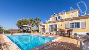 A swimming pool with a tiled deck, a covered seating area, and a building with a tiled roof and windows.