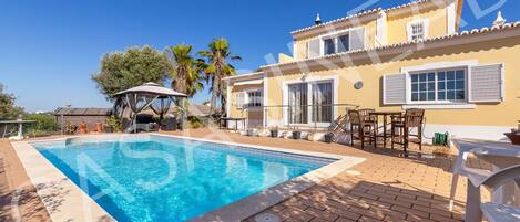 A swimming pool with a tiled deck, a covered seating area, and a building with a tiled roof and windows.