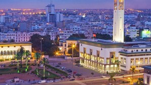 Une vue urbaine avec un beffroi imposant, une fontaine centrale et des bâtiments environnants.