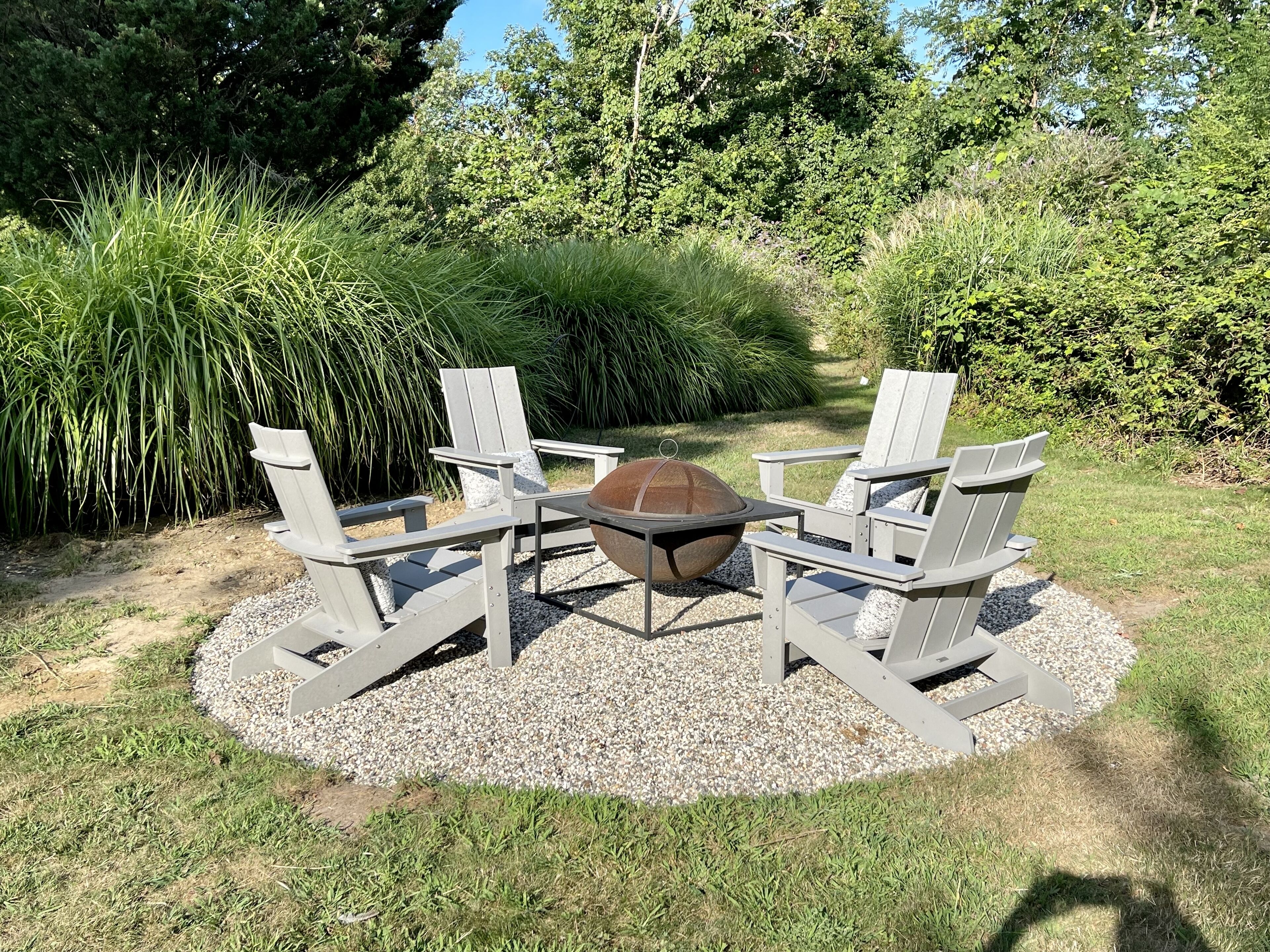 A fire pit surrounded by four Adirondack chairs in a garden setting.