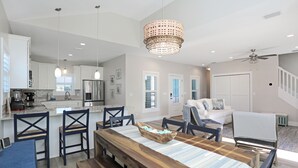 Un'ampia zona giorno con tavolo da pranzo, sedie e un lampadario. Si può vedere una cucina sullo sfondo, con mobili bianchi e un frigorifero.