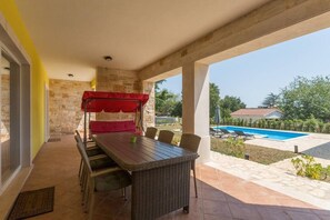 Um pátio externo com mesa e cadeiras, área de estar coberta e vista para uma piscina e vegetação.