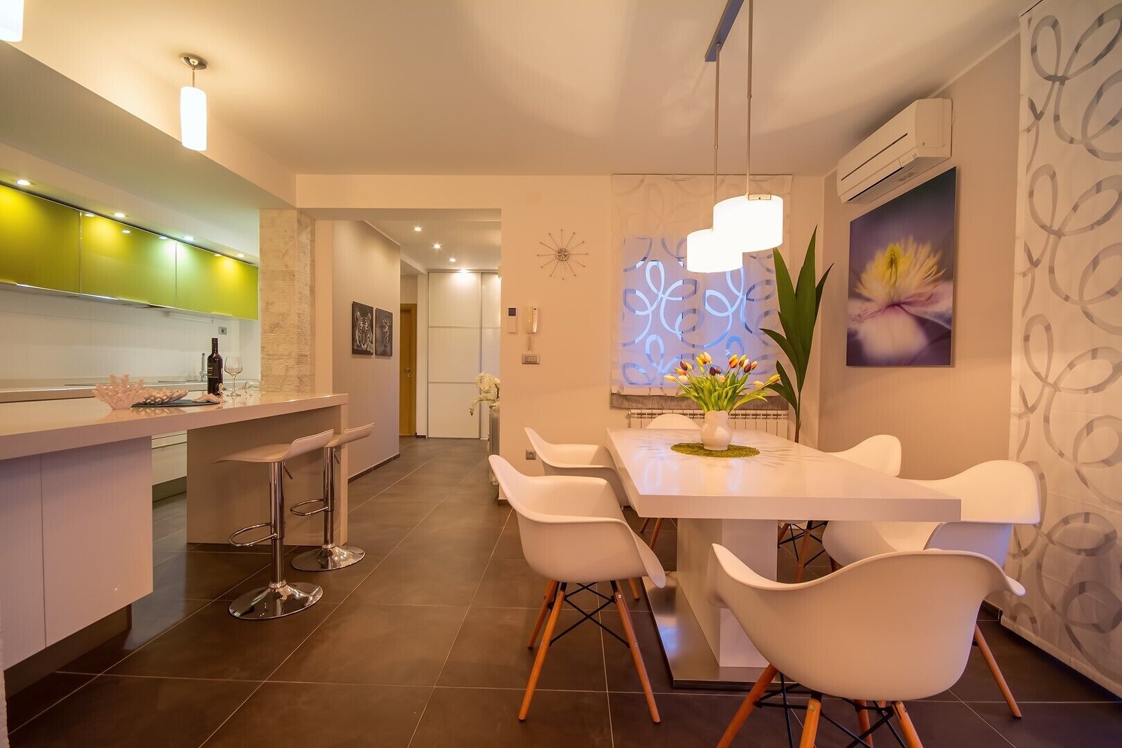 A modern kitchen with a central island, white bar stools, and a dining area with white chairs and a vase of flowers.