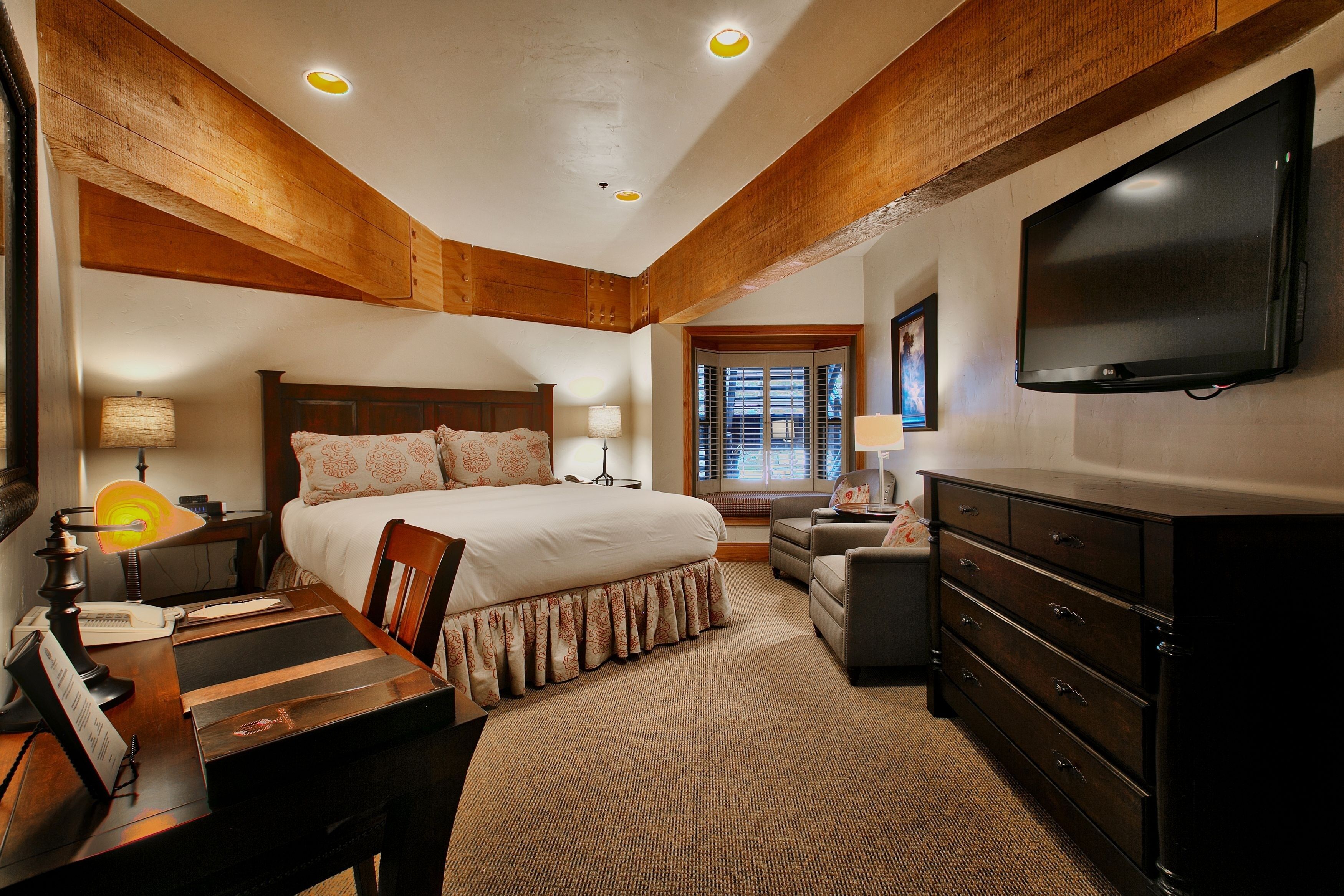 A bedroom with a large bed, wooden ceiling beams, a flat-screen TV, a desk with a lamp, and a dresser.