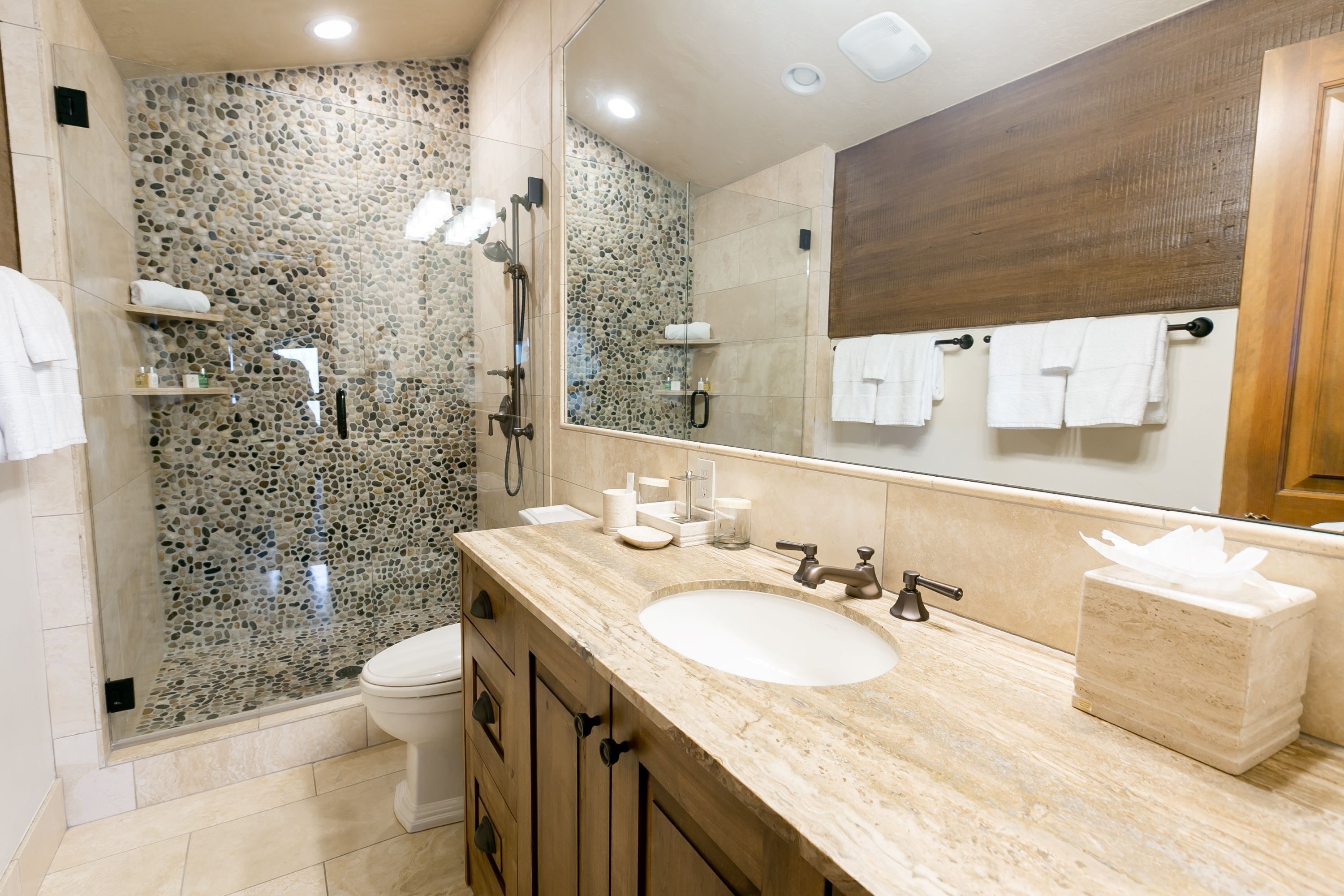 A modern bathroom with a stone shower wall, a large mirror, a sink with a marble countertop, and a toilet.