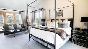 Una cama con dosel con un edredón blanco y marrón, estructura de metal negro y varias almohadas. Hay un sofá de cuero negro con cojines blancos y una mesita con una lámpara. La habitación tiene ventanas grandes con cortinas, un cuadro enmarcado en la pared y una mesita de noche de metal negro con una lámpara.