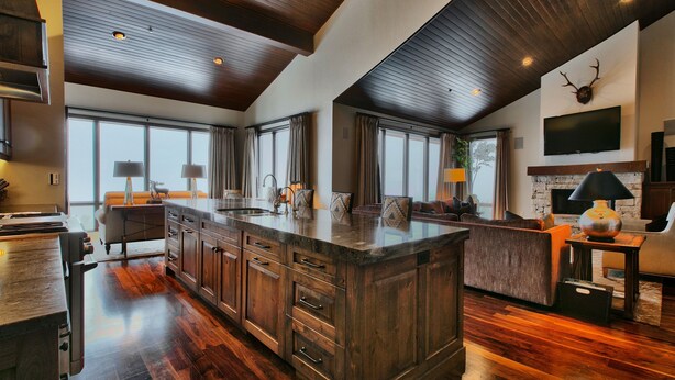 A spacious kitchen with a large island, wooden cabinets, and a stone backsplash. There is a built-in oven, a sink, and a dining area with a table and chairs.