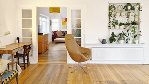 A modern living room with a wooden floor, a brown chair, a dining table with chairs, a white radiator, and a plant shelf. - Coastal - Wittering  -  an annexe that sleeps 6 guests  in 3 bedrooms (West Wittering)