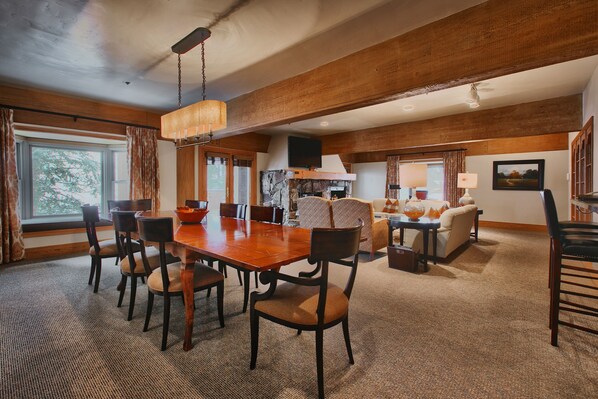 A spacious dining area with a wooden table and chairs, a kitchen in the background, and a large window with curtains.