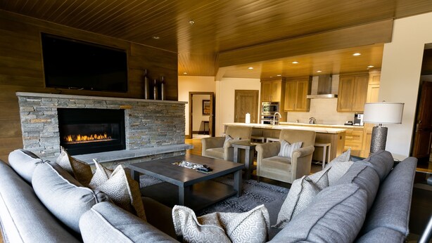 A modern living room with a stone fireplace, a large gray sofa, and a kitchen area visible through an open doorway. - Luxury Two Bedroom Corner Suite with Mountain Views 2 Apartment Hotel by RedAwning (Park City)