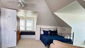 A bedroom with a sloped ceiling, a bed with a metal frame, a beige sofa, and a door leading to another room. - Beachfront Home with Sunset Views, Steps to Bay (Cape Charles)
