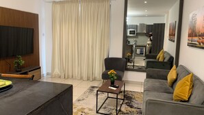 Uma sala de estar moderna com sofá, mesa de centro e televisão. Uma cozinha visível por uma porta aberta.