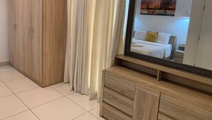 Quarto com guarda-roupa de madeira, espelho, cama com roupa de cama branca e um quadro na parede.