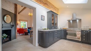 A kitchen with dark grey cabinets, a built-in oven, and a chimney. There is a fireplace with a mirror above it. The kitchen connects to a dining area with red chairs.