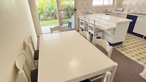 Une cuisine avec une table et des chaises blanches, un évier et une fenêtre donnant sur un jardin.