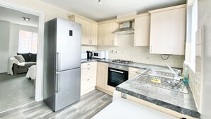 A modern kitchen with a stainless steel refrigerator, a microwave, a gas stove, and a dishwasher. There is a window with a view of a building outside, and a sofa in the adjacent living area. - Ammaculate 3 Bedroom Property Near Caltic Manor (Newport)