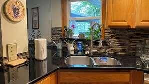 A kitchen with wooden cabinets, a black countertop, a stainless steel sink, and a window with a view of a house and trees. - Seneca Riverfront Spa! Hot tub, Pool + Sauna! Pontoon Boat and Canoe Available! (Baldwinsville)