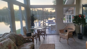 A sunlit patio with wicker furniture, a glass door leading to a deck, and a view of a lake. - Seneca Riverfront Spa! Hot tub, Pool + Sauna! Pontoon Boat and Canoe Available! (Baldwinsville)